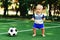 Little boy in blue shorts playing with soccer ball at sports ground. Blonde child in uniform standing at football field with ball