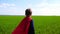 A little boy dressed in a superman costume runs across a green field on a sunny day.