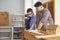 Little boy drills holes in a wooden board under the control of his dad in a home workshop.