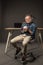 little boy in eyeglasses holding coffee cup and sitting on chair near table with laptop and lamp on grey