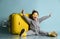 Little boy in gray overall, yellow sunglasses, rubber boots. Sitting on floor, suitcase, raised hands, posing on blue background