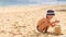 Little Boy in Hat Makes Sand Cakes with Large Plastic Glass