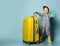 Little boy kid in gray overall in yellow sunglasses and rubber boots holding suitcase luggage posing on pastel blue studio