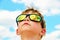 A little boy looks at the cloudy sky with glasses. The clouds are reflected in the glass. Yellow glasses against the blue sky.
