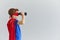 Little boy in a mask and cloak of a superhero looks through binoculars on a gray background.