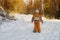 Little boy in an orange jumpsuit standing on snow-covered road in a coniferous forest. Mother is in a distance. Winter sunny day.