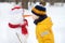 Little boy playing with funny snowman. Child reaches for a snowman`s carrot nose and wants to bite. Active outdoors leisure with