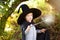 Little boy in pointed hat and black cloak playing with magic wand outdoors. Little wizard