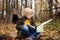 Little boy scout is orienteering in forest. Child is sitting on fallen tree and looking on map on background of teepee hut