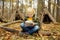 Little boy scout is orienteering in forest. Child is sitting on fallen tree and looking on map on background of teepee hut.