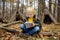 Little boy scout is orienteering in forest. Child is sitting on fallen tree and writing in the notepad. Behind the child is teepee