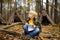 Little boy scout is orienteering in forest. Child is sitting on fallen tree and writing in the notepad. Behind the child is teepee