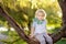 Little boy is sitting on a branch of big tree and is dreaming. Child`s games. Active family time on nature. Hiking with kids