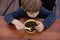 Little boy sitting on chair at brown wooden table in kitchen and having breakfast top view. Kid of kindergarten age