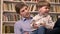 Little boy sitting with his young father and holding ball, shelves with books background