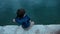 Little boy sitting on pier throws stones into water outdoors.