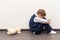 A little boy of six is offended and sits on the floor against a white wall next to a soft toy with his hands closed