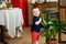 A little boy stands in the middle of a room next to a large indoor plant