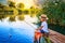 Little boy in straw hat sitting on the edge of a wooden dock and fishing in lake at sunset