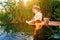 Little boy in straw hat sitting on the edge of a wooden dock and fishing in lake at sunset