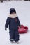 Little boy in warm coat and hat pulling his sledge in cold winter day. It`s snowing