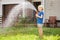 Little boy watering lawn and playing with garden hose with sprinkler in sunny backyard. Preschooler child having fun with spray of