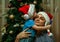 A little boy wearing Santa Claus hat and his uncle wearing Snow Maiden hat sitting on background of New Year tree.