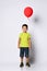 Little brunet boy in yellow t-shirt, denim shorts and khaki sneakers. Smiling, holding red balloon, posing isolated on white