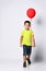 Little brunet boy in yellow t-shirt, denim shorts and khaki sneakers. Smiling, holding red balloon, posing isolated on white