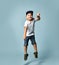 Little brunet child in white t-shirt, denim shorts and khaki sneakers. Jumping up, holding gray cap, posing on blue background