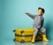 Little brunet kid in gray overall, yellow sunglasses, rubber boots. He sitting on suitcase, pointing, posing on blue background