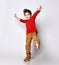 Little brunet kid in red jumper, brown pants and sneakers. He smiling, raised his hands and leg up, posing isolated on white