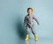 Little brunet model in orange hat, gray overall and yellow rubber boots. He is smiling while posing against blue studio background