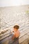 Little Caucasian boy child sitting on wooden pier sandy beach, summer time, sea vacation near water. The theme is the flow of time