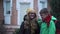 Little Caucasian boy in green coat hugging pretty girl in eyeglasses and yellow hat next to the big house. Positive