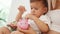 Little caucasian brunette boy sitting on his mother's lap and carefully putting coins in his pink piggy bank