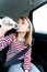 Little caucasian girl is driving in car, dreanking pure water from plastic kids bottle. Traveling on road in safe baby seats with