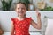 Little caucasian girl in red dress sitting on sofa, smiling with happy face doing victory sign