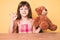 Little caucasian kid girl with long hair sitting on the table with teddy bear doing ok sign with fingers, smiling friendly