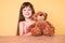 Little caucasian kid girl with long hair sitting on the table with teddy bear looking positive and happy standing and smiling with
