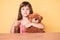 Little caucasian kid girl with long hair sitting on the table with teddy bear smiling happy and positive, thumb up doing excellent