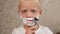 A little cheerful boy with shaving foam on his face shaves.