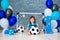 Little cheerful child dressed in sports clothes sitting on the floor near a football goal, looking at a big soccer ball. The first