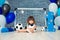 Little cheerful child dressed in sports clothes sitting on the floor near a football goal, looking at a big soccer ball. The first