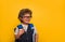 Little child boy with book and bag isolated on yellow paper wall. Happy smiling kid go back to school, kindergarten