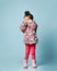Little child dressed in pink faux fur coat, pants and boots. She covered eyes with hands, posing on blue background. Full length