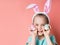 Little child in headband with rabbit ears, blue t-shirt. She smiling, showing two colorful eggs, posing on pink