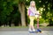 Little child learning to ride a scooter in a city park on sunny summer day. Cute preschooler girl in safety helmet riding a roller