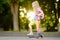 Little child learning to ride a scooter in a city park on sunny summer day. Cute preschooler girl in safety helmet riding a roller