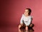 Little child in smart white bodysuit with inscription, barefoot. She smiling, sitting on floor against pink background. Close up